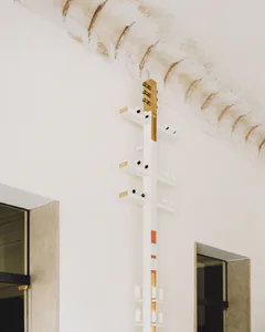 Vertical sculptural wall light fixture made of white shelves, brass accents, and electric candles mounted on a white plaster wall under a curved, aged vaulted ceiling with two doorways visible on either side.