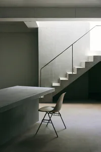 Minimalist brutalist interior with raw concrete walls, a floating concrete staircase with thin metal handrail, and a beige Eames-style chair placed next to a concrete counter.