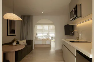 Modern minimalist studio apartment interior with white arched doorway leading to bedroom, green sofa, round wooden table, and compact kitchen with built-in microwave and induction cooktop.