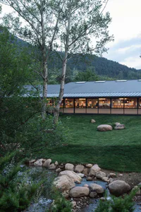 Modern single-story mountain house with large glass windows and corten steel roof, overlooking a rocky stream and lush lawn at dusk.