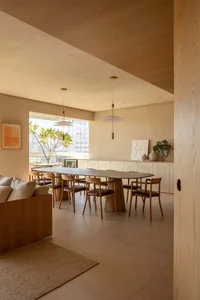 Modern minimalist dining room featuring a large oval travertine-topped oak dining table surrounded by wooden chairs, two white pendant lights, built-in oak cabinetry, and a large window overlooking city buildings.