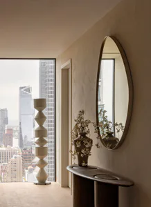 Modern minimalist entryway in a New York high-rise apartment featuring a tall sculptural beige floor lamp, an oval mirror, a dark organic-shaped travertine console table with a vase of white cherry blossoms, and a panoramic city view through large windows.