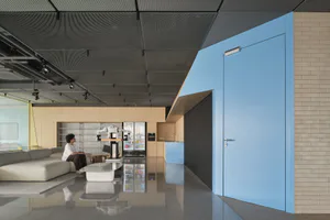 Modern open-plan office lounge featuring a large white robotic arm, reflective polished concrete floor, light gray sectional sofa, blue door, and beverage refrigerators.
