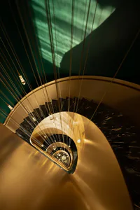 Overhead view of a luxurious spiral staircase featuring black marble treads, gold metallic structure, and dramatic pendant lighting against a teal background.