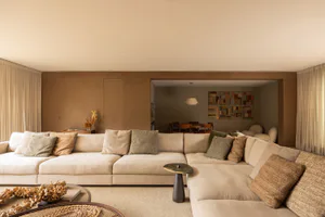 Modern minimalist living room with a large L-shaped sectional sofa in neutral beige fabric, multiple textured throw pillows in beige, green and brown tones, round coffee tables with natural woven and wood elements, light wood panel walls, and a view through to a dining area with colorful patchwork wall art.