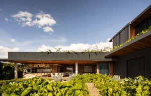 Modern tropical house exterior featuring a large covered outdoor patio with wooden ceiling, woven pendant lights, stone walls, and lush green tropical plants in the foreground under a bright blue sky.