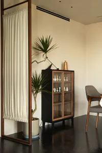 Modern minimalist living room corner with a black and walnut wood display cabinet filled with ceramic vases, a large potted yucca tree, a wooden room divider with white curtain, and a grey upholstered wooden armchair on dark reflective flooring.