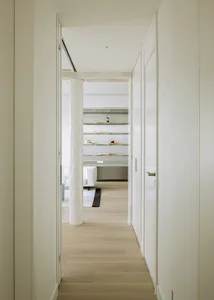 View from a narrow white hallway looking into a modern study area featuring a cylindrical mother-of-pearl column, built-in floating shelves with decorative objects, light wood flooring, and a white sofa in the background.