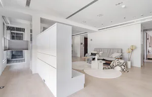 Modern minimalist living room featuring a tall white lacquered storage partition, light wood flooring, two overlapping circular beige rugs, a grey fabric sofa, and a small round coffee table with white flowers.
