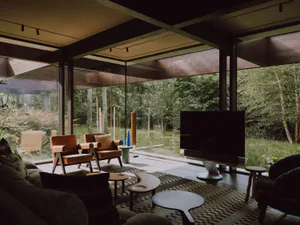Modern living room interior featuring large glass walls overlooking a forest, orange leather armchairs, wooden ceiling beams, a large flat-screen TV, and a patterned area rug with organic-shaped coffee tables.