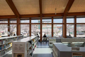 Modern library interior with long white bookshelves, wooden ceiling beams, large windows overlooking cherry blossom trees in full bloom, and people quietly reading.