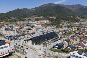 Aerial view of a modern solar community center with a large black photovoltaic glass roof, surrounded by parking lots, roads, and residential buildings with cherry blossoms in full bloom against a forested mountain background.