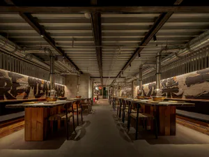 Interior view of a modern industrial-style hotpot restaurant featuring long reclaimed wood tables, concrete floors, exposed metal ductwork, and graffiti-style mural walls.