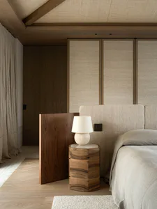 Modern minimalist bedroom featuring a curved walnut wood nightstand with a travertine table lamp, beige upholstered headboard, wooden ceiling beams, and neutral linen bedding.