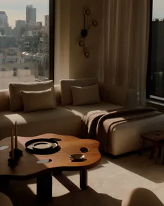 A modern minimalist living room featuring a beige L-shaped sectional sofa, a sculptural walnut amoeba-shaped coffee table with black trays and candle holders, a brown throw blanket, and a unique wall sconce, with large windows showing a city view.
