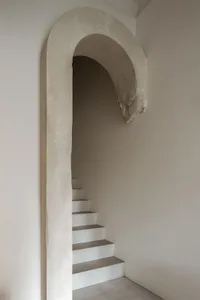 View of a minimalist concrete staircase ascending through a large, hand-finished arched opening in a white plastered wall.