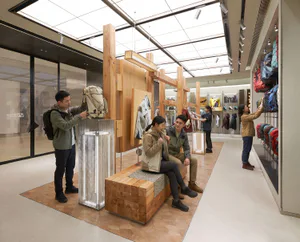 Modern retail interior of an outdoor gear store featuring a large modular wooden display structure with metal mesh lighting columns and customers browsing merchandise.