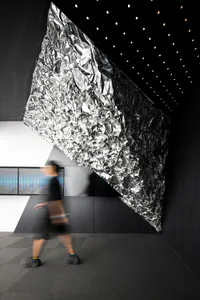 Modern art installation featuring a large crumpled metallic foil structure hanging diagonally in a dark exhibition space with dotted ceiling lights, a person walking by creating motion blur.