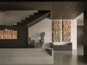 Modern entrance hall featuring a grand stone staircase, dark wood ceiling, built-in display of tribal wooden masks, two large wall sculptures made of stacked masks, and a sculptural black bench under dramatic lighting.