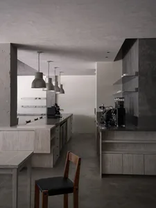 Modern minimalist coffee bar interior with raw concrete walls, light wood cabinetry, black espresso machines, and a row of industrial-style metal pendant lights hanging over a long counter.