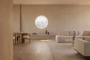 Modern minimalist living room interior featuring light oak flooring and furniture, a large circular window with city view, a low-profile beige sectional sofa, and a built-in window bench with decorative objects.