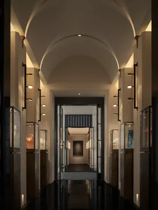 Long symmetrical hallway in a contemporary art gallery with vaulted arched ceiling, polished dark marble floor, and illuminated glass display cases containing sculptural art pieces.