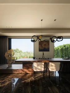 Contemporary dining room featuring a long dark wood table, two white upholstered chairs, a large abstract sculptural chandelier, a golden geometric sculpture on a raised platform, and large windows overlooking green hills.