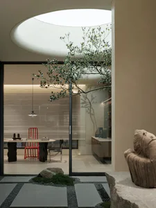 Modern minimalist dining room viewed through glass doors from a Japanese-style courtyard featuring stone pavers, gravel, moss, a large rock, and a carved stone statue, with a red chair and organic dark wood table visible inside under a circular skylight.