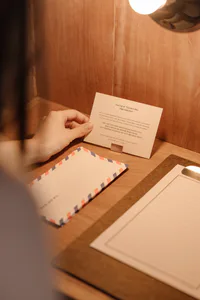 一隻手在溫暖的胡桃木書桌上拿著類比打字機說明卡，旁邊有航空郵件信封與橫線信紙，沐浴在柔和燈光下。