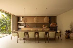 Modern open-plan dining room with a long dark wood table, ten upholstered chairs, three large woven rattan pendant lights, a built-in kitchen with wooden cabinetry, and a copper pizza oven visible in the background, opening to a lush tropical garden.