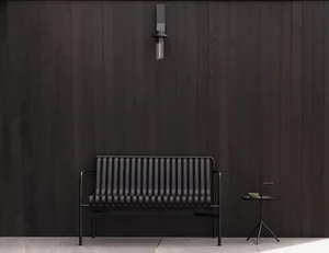 Modern black metal outdoor sofa with vertical slats and a small round side table holding a green cup, placed against a dark vertical wood-paneled wall with a black wall sconce above.