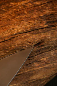 Close-up photograph of a metallic knife blade inserted into a dark, weathered walnut wooden surface with prominent grain and cracks.