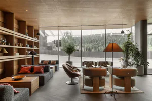 Modern interior lounge featuring built-in oak shelving, grey modular sofas with orange cushions, brown leather swivel chairs, and large floor-to-ceiling windows overlooking a dense green forest.