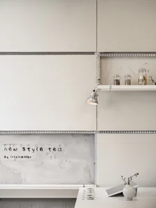 Minimalist kitchen corner featuring a large concrete wall with handwritten text 'new style tea by inspiration', a white floating shelf holding glass jars, a silver industrial-style wall lamp, and a white countertop with a small vase of dried branches and stacked coasters.