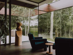 Modern living room interior featuring two teal upholstered armchairs, a small round wooden side table, a large abstract wooden sculpture on a pedestal, and expansive glass walls overlooking a lush green forest with birch trees.