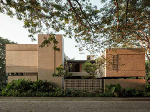 Modern tropical house featuring exposed concrete walls, red brick volumes, wooden lattice screens, and abundant tropical landscaping under a clear sky.