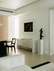 Modern minimalist living and dining area featuring a white shagreen console table, black abstract sculpture on a pedestal, framed dark portrait painting, round dark wood dining table with black woven-back chairs, and light wood flooring.