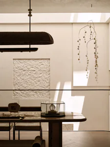 Modern minimalist dining room interior with a large travertine textured wall panel, a dark sculptural pendant lamp, a delicate metal leaf mobile hanging from the ceiling, a marble dining table with a glass display case, and woven dining chairs.