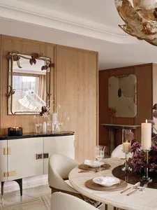 Elegant contemporary dining room featuring light oak cabinetry, a sculptural brass mirror, marble dining table, and cream upholstered chairs under a dramatic floral chandelier.