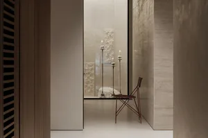 Minimalist interior view of a travertine-clad alcove featuring three tall candlesticks with lit candles, a large rough stone sculpture, and a distinctive red wireframe folding chair positioned against textured stone walls.