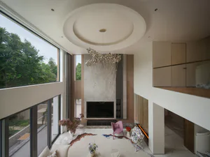 Modern double-height living room interior with large circular ceiling detail, sculptural chandelier, marble flooring, pink accent chair, and floor-to-ceiling windows showing trees outside.