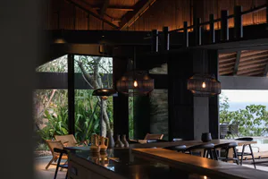 Modern open-plan dining area featuring large woven rattan pendant lights, dark wood beams, a long wooden dining table with black stools, rattan lounge chairs, and expansive glass walls opening to tropical vegetation and an ocean view.