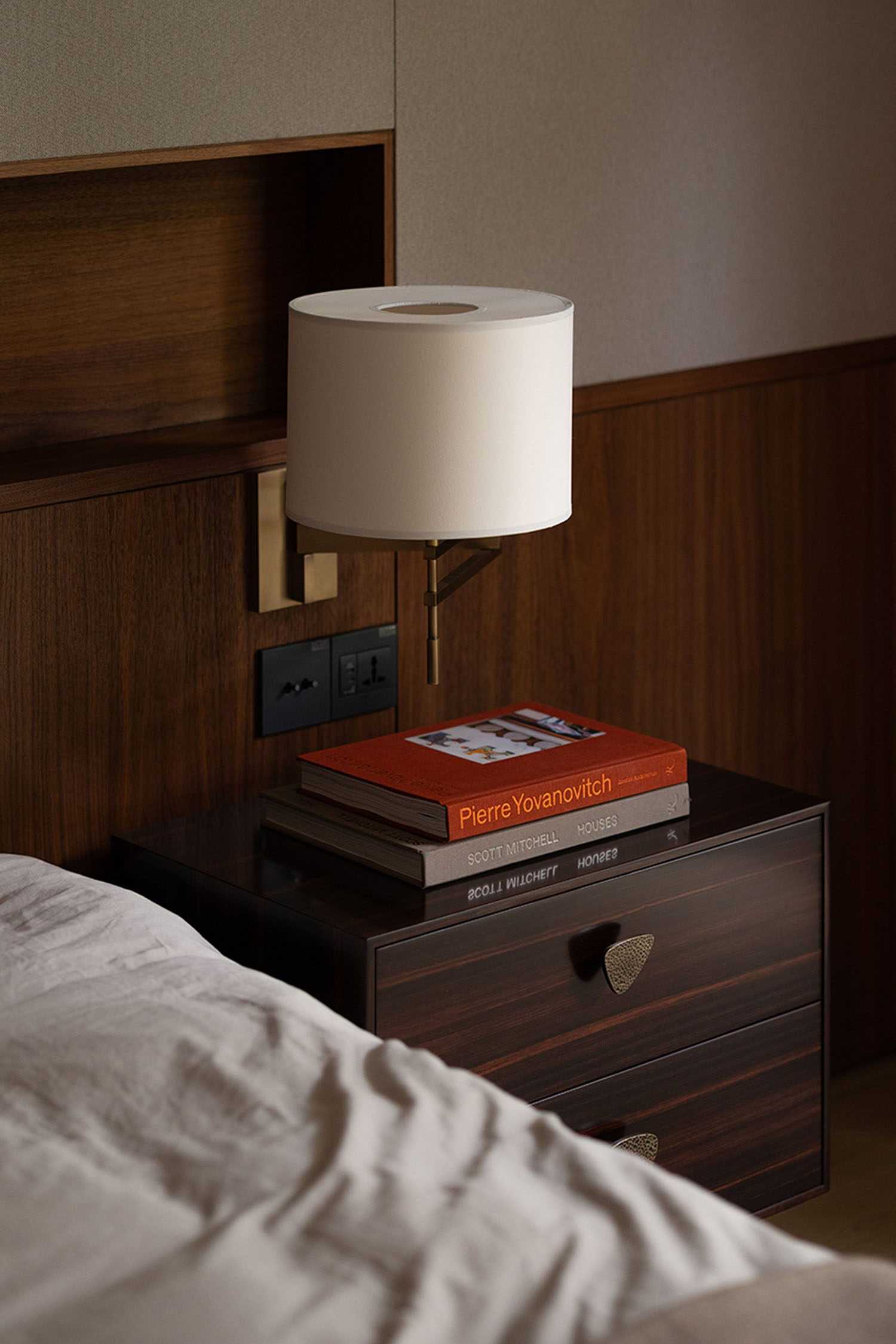 Dark wooden bedside drawers with books and a shaded wall lamp beside a bed.