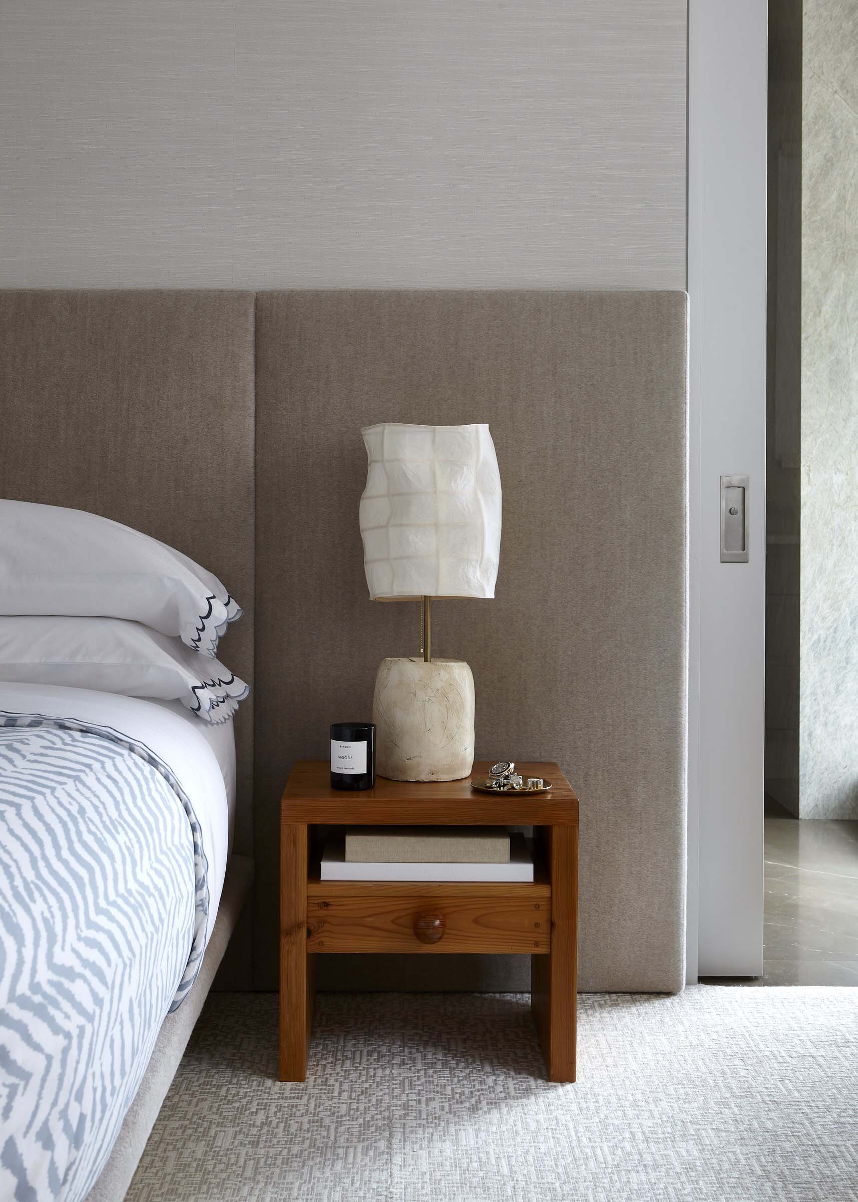 Wooden bedside table with a drawer, open shelf, and shaded lamp beside a bed.