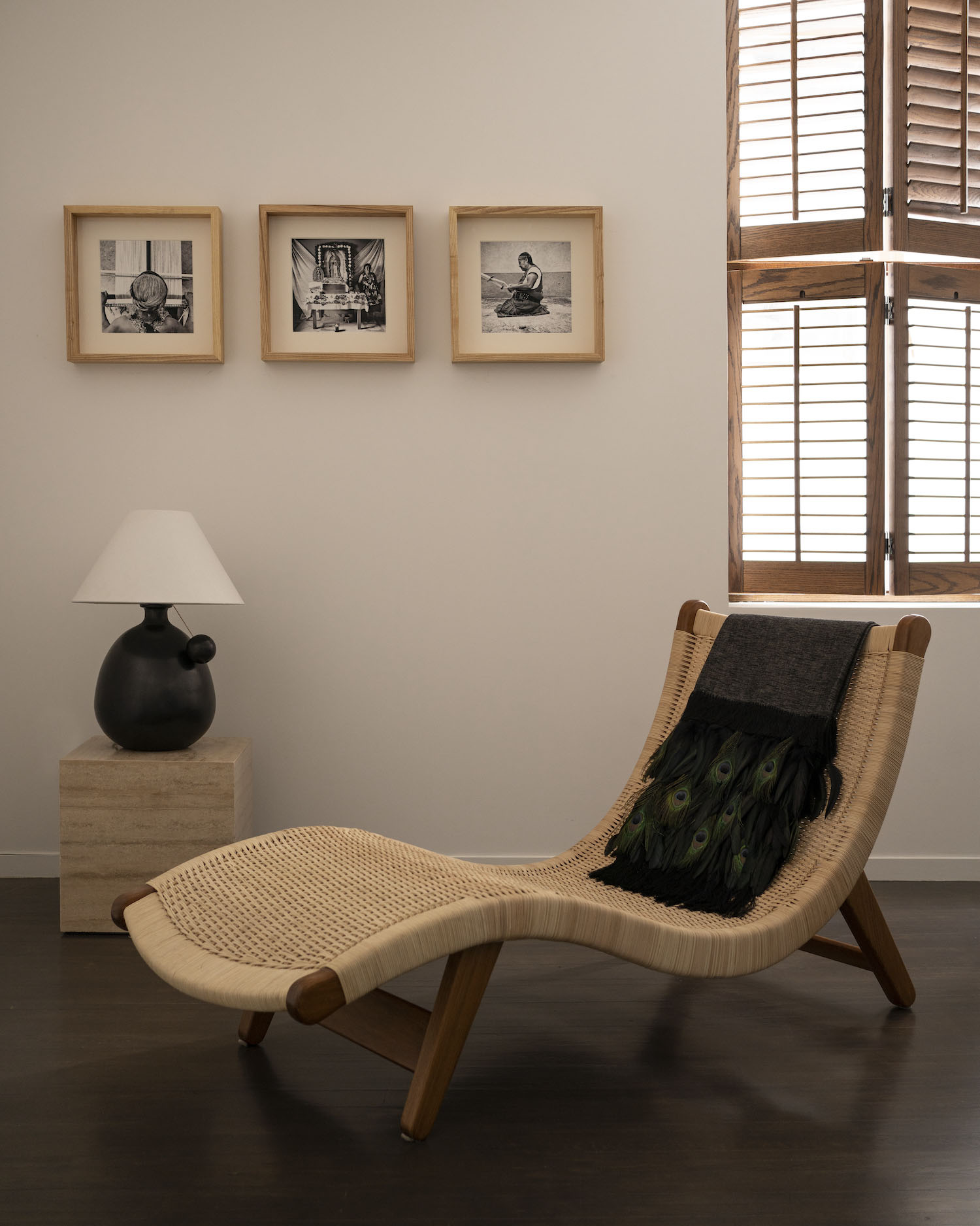 Warm neutral lounge corner with a woven chaise, table lamp, and wooden window shutters.