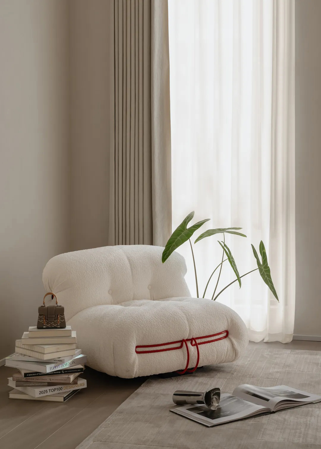 Soft white lounge chair with red trim beside sheer curtains and a stack of books.